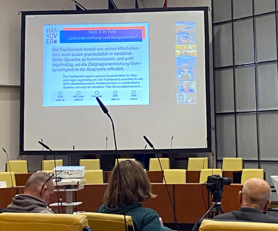 Im Vordergrund drei Menschen die auf gelb gepolsterten St&uuml;hlen sitzen und der Kamera den R&uuml;cken zuwenden. Gro&szlig; im Bild eine Leinwand mit einer Pr&auml;sentation.