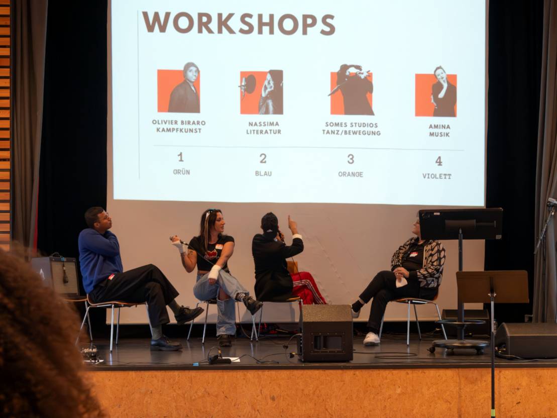 Vier Personen sitzen auf einer Bühne. Im Hintergrund eine Leinwand, auf die eine der Personen zeigt. Auf der Leinwand steht "Workshops", darunter sind vier Bilder von anderen Personen.