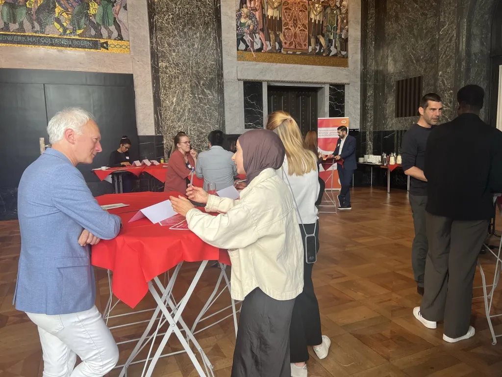 Zwei Menschen stehen an einem Stehtisch mit Roter Tischdecke. Auf der linken Seite ein &auml;lterer Mann mit wei&szlig;en Haaren und blauem Sakko, auf der rechten Seite eine junge Frau mit beiger Jacke und Kopftuch. Sie sprechen miteinander. Im Hintergrund weitere Stehtische und Menschen.