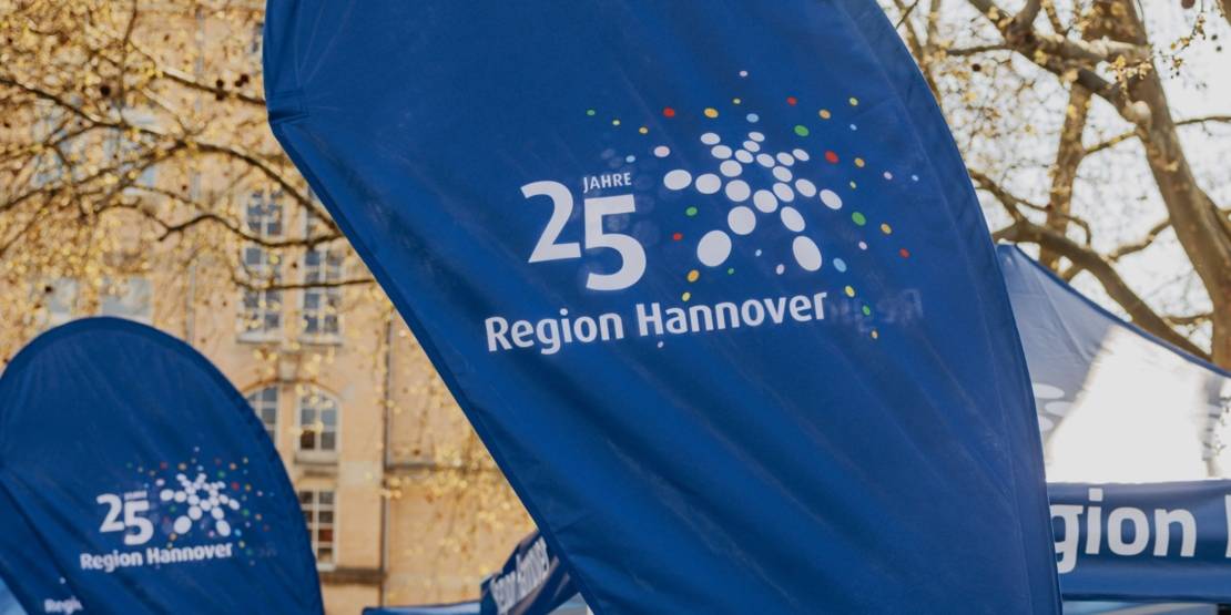 Eine Beachflag in der Mitte des Bilds mit Aufschrift "25 Jahre Region Hannover", im Hintergrund Pavillons und weitere Beachflags.