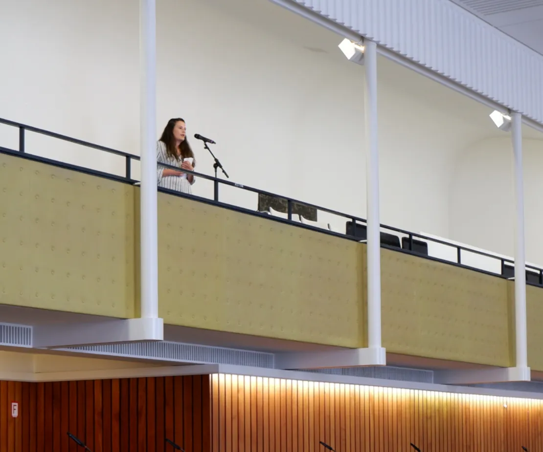 Eine Frau steht auf der ZuschauerInnen-Trib&uuml;ne des Ratssaals und spricht in eine Mikrofon. In der Hand h&auml;lt sie einen Zettel.