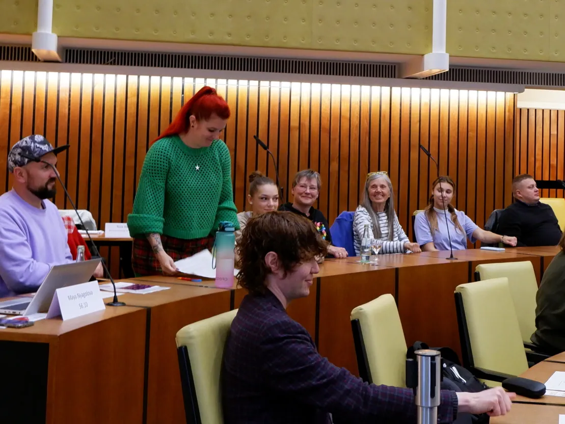 Eine Frau im gr&uuml;nen Strickpullover steht vor einem Tischmikrofon und sortiert Unterlagen. Mehrere Personen sitzen neben ihr und schauen sie l&auml;chelnd an.