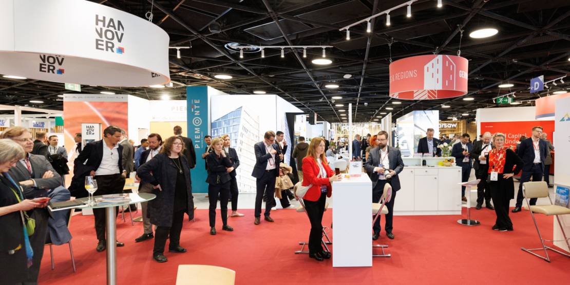 Menschen stehen in einer Halle mit Stehtischen und Ständen.