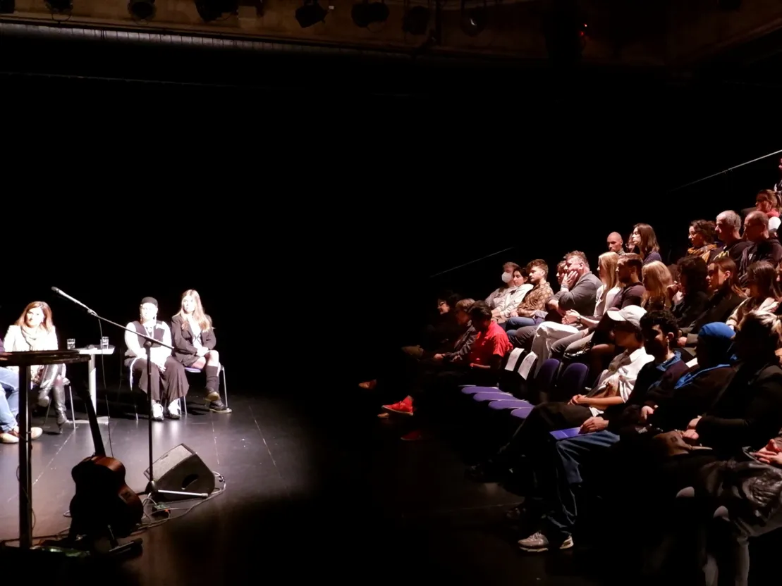 Linke Bildh&auml;lfte: Personen, die in einer Diskussionsrunde sitzen. Rechte Bildh&auml;lfte: Ein Publikum auf einer Trib&uuml;ne. Die Szene findet in einem sehr dunklen Raum statt. Die Podiumsteilnehmer*innen sind stark ausgeleuchtet, das Publikum ist schwach ausgeleuchtet.