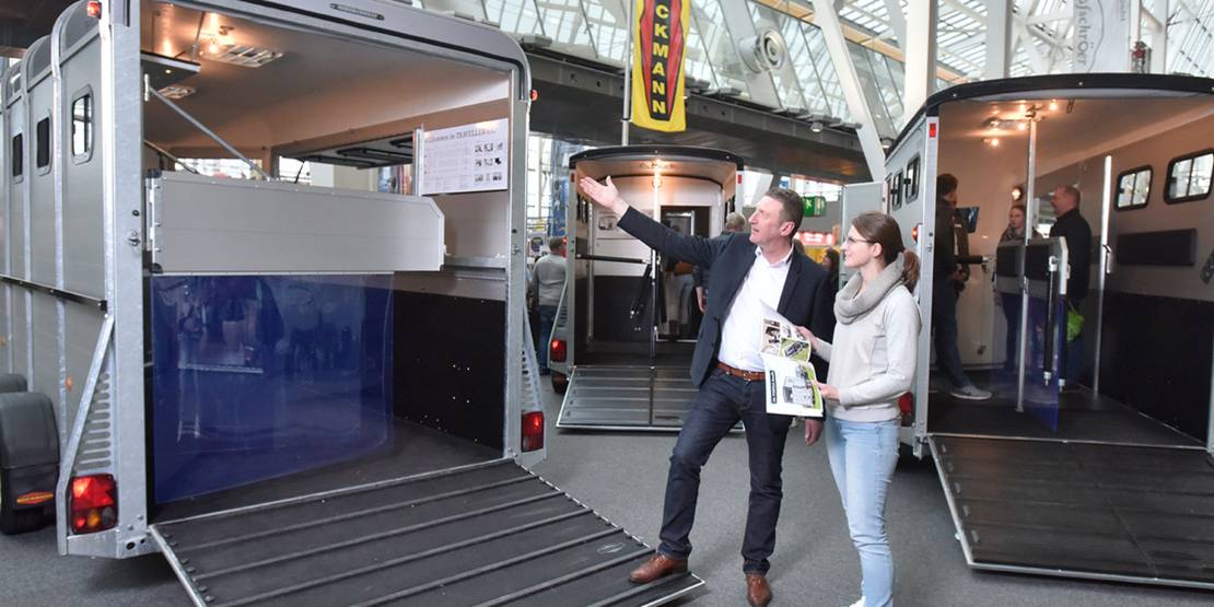 Ein Mann präsentiert einer Frau einen geöffneten Pferdeanhänger.