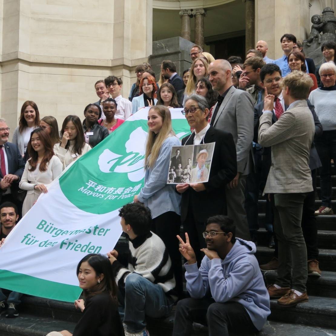 Die Teilnehmenden der Jugendfriedenskonferenz präsentieren die Fahne von Mayors for Peace auf der Rathaustreppe.