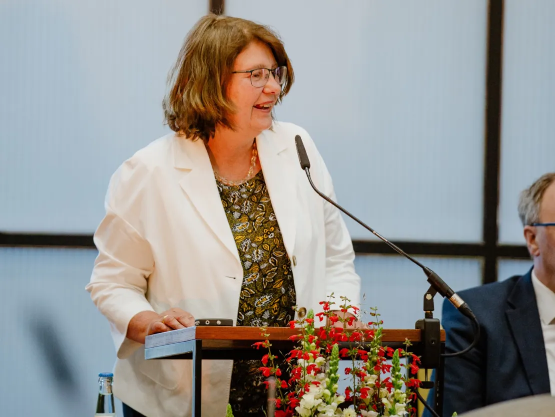 Eine Frau in wei&szlig;em Blazer steht an einem Redepult. Sie tr&auml;gt eine Brille und l&auml;chelt. Im Vordergrund sind vor dem Pult Blumen zu sehen.