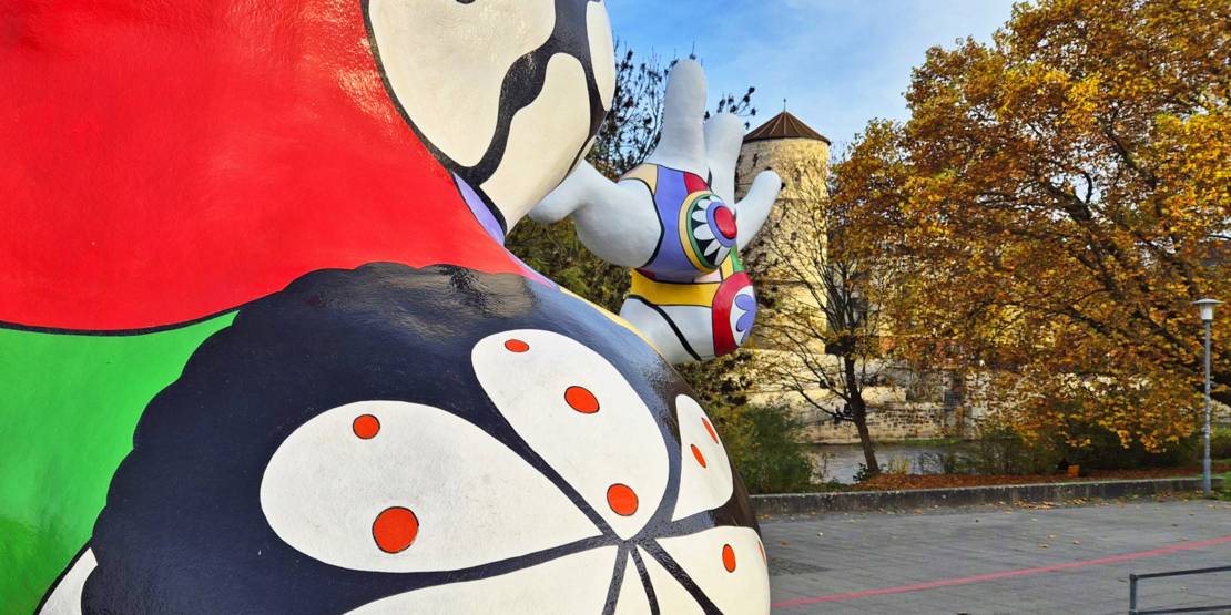 Die Nanas vor herbstlich belaubten Bäumen, im Hintergrund der Beginenturm.