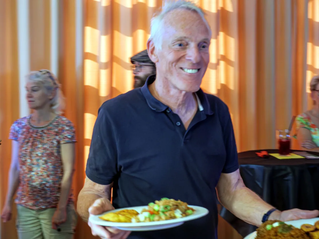 Ein breit grinsender Mann mit wei&szlig;en Haaren tr&auml;gt zwei Teller mit Essen. Er tr&auml;gt ein blaues Poloshirt und sieht so aus, als w&uuml;rde er das Essen servieren.