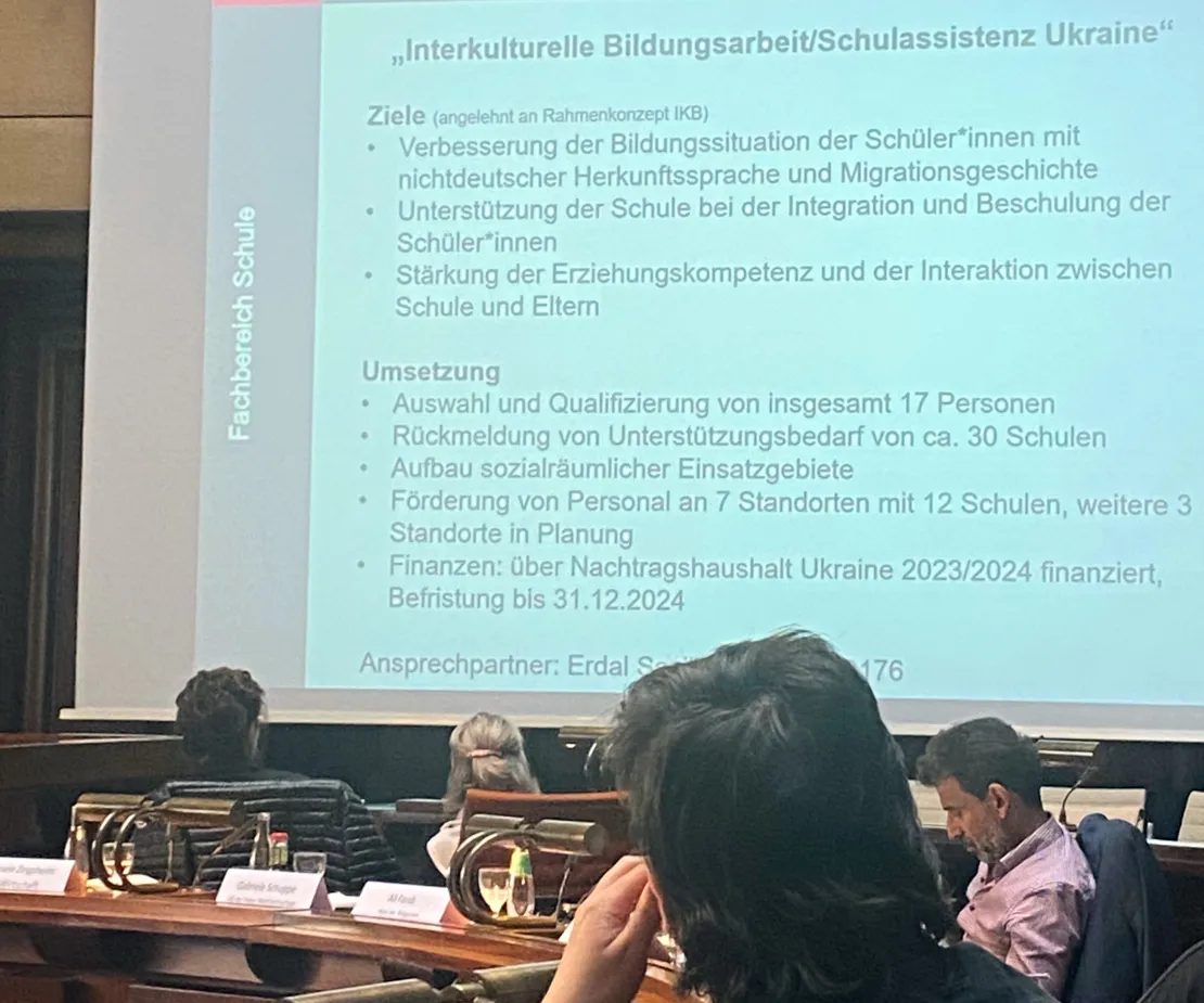 Eine Folie auf einer Leinwand nimmt fast das gesamt Bild ein. &Uuml;berschrift der Folie ist "Interkulturelle bildungsarbeit/Schulassistenz Ukraine". Im Vordergrund die K&ouml;pfe von vier Personen. Drei davon schauen zu der Leinwand.