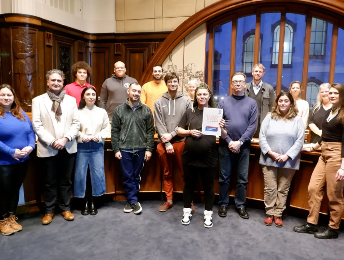 16 Personen stehen f&uuml;r ein Gruppenfoto vor einer halbrunden Plenar-Tisch-Reihe. Sie l&auml;cheln in die Kamera. Eine Frau in der Bildmitte h&auml;lt den WIR2.0-Ma&szlig;nahmenkatalog in die Kamera. Im Hintergrund ein gro&szlig;es Fenster in einen Innenhof des Neuen Rathauses.