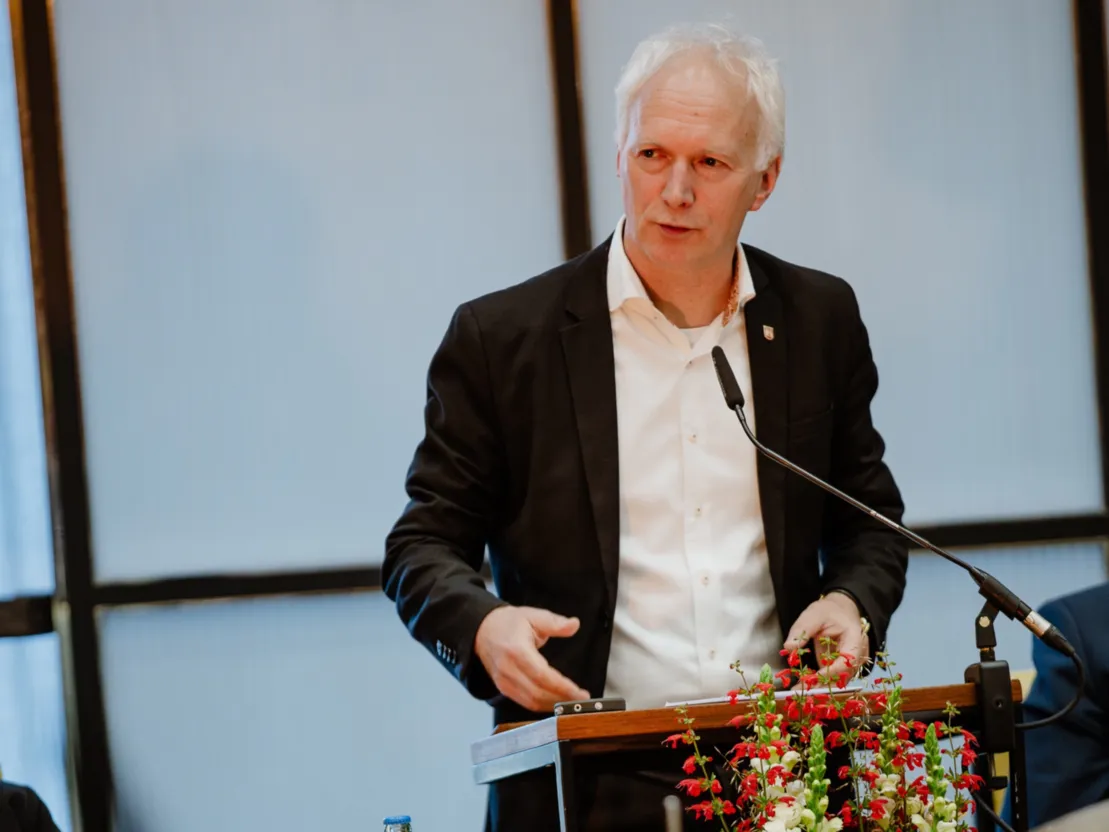 Ein Mann in schwarzem Sakko und wei&szlig;em Hemd steht an einem Redepult in der rechten Bildh&auml;lfte. Er schaut nach links vorne.