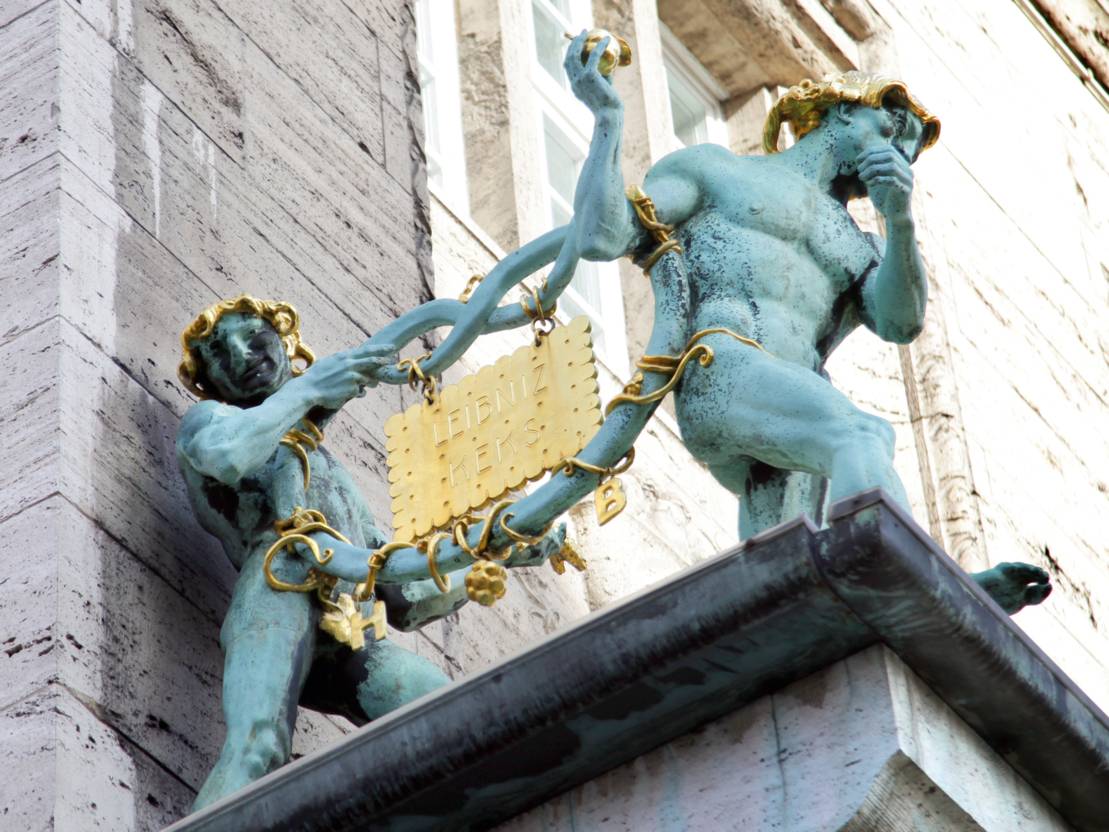 Zwei bronzene Figuren, die zusammen eine ebenfalls Bronzene Brezel tragen. In diese Brezel ist ein goldener Keks eingeh&auml;ngt - er hat die Aufschrift "Leibniz Keks".