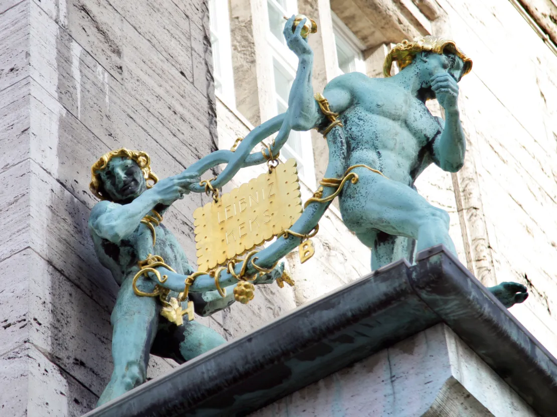 Zwei bronzene Figuren, die zusammen eine ebenfalls Bronzene Brezel tragen. In diese Brezel ist ein goldener Keks eingeh&auml;ngt - er hat die Aufschrift "Leibniz Keks".