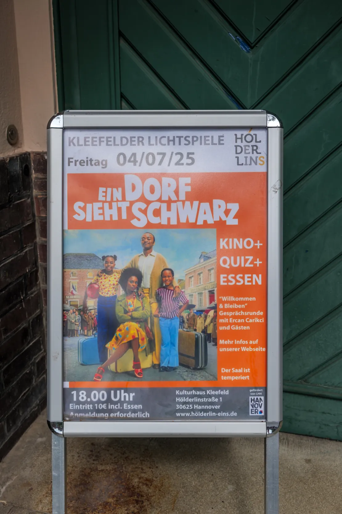 Ein Filmplakat der franz&ouml;sischen Kom&ouml;die "Ein Dorf sieht Schwarz" in einem Aufsteller vor einer gr&uuml;nen T&uuml;r. Auf dem Filmplakat ist eine schwarze Familie mit Mutter, Vater und zwei Kindern zu sehen. Im Hintergrund des Plakats eine durchweg wei&szlig;e Dorfgesellschaft.