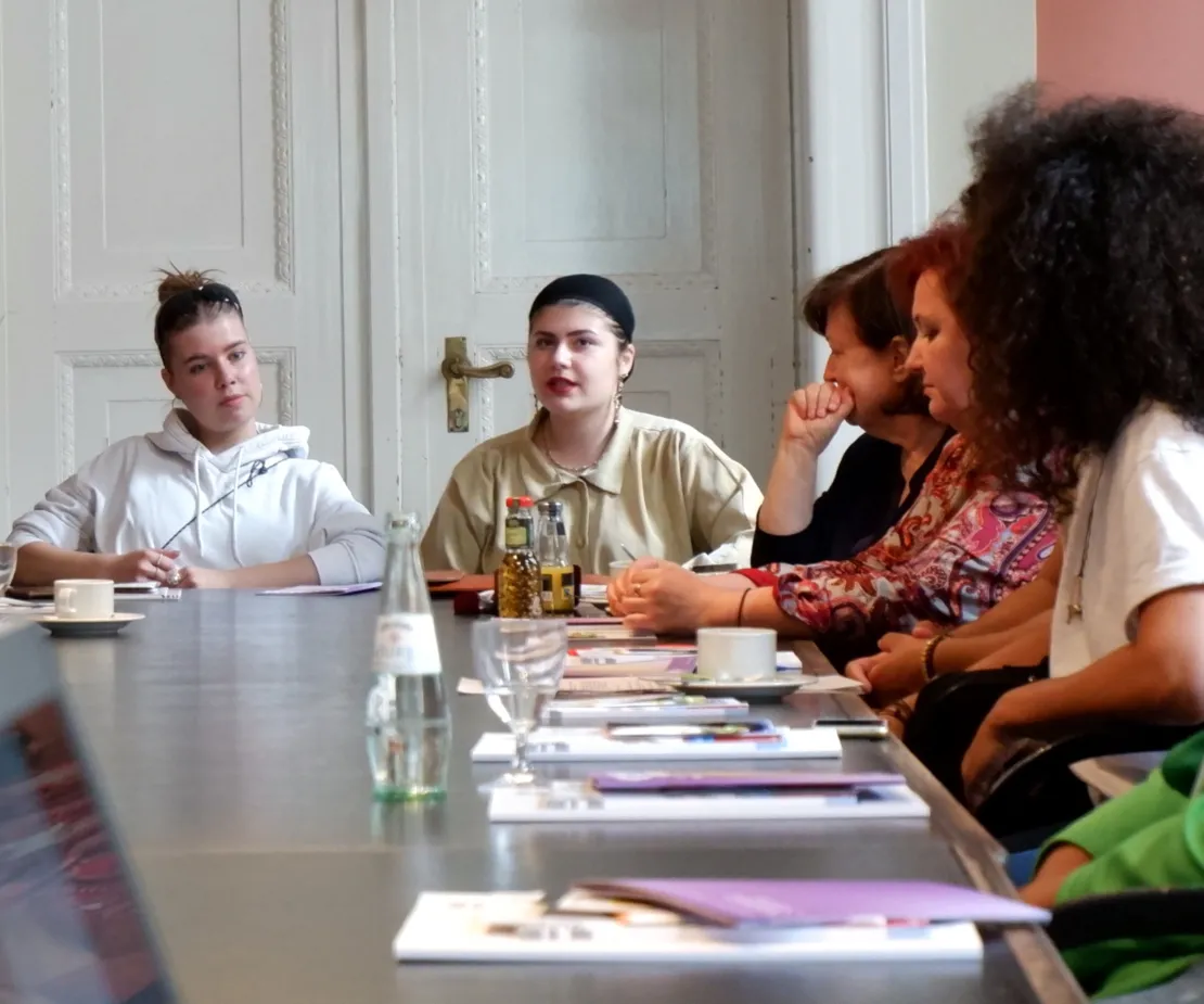 Zwei junge Frauen sitzen vor dem Ende eines langen Tisches. Auf der rechten Bildseite sieht man mehrere Menschen an der langen Seite des Tisches sitzen. Sie h&ouml;ren den Frauen zu.