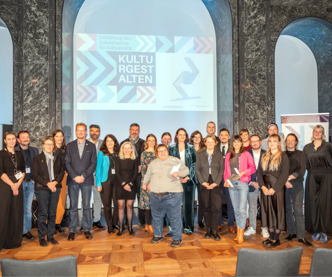 24 Menschen posieren f&uuml;r ein Gruppenfoto und l&auml;cheln in die Kamera. Einige von ihnen halten Preis-Skulpturen in der Hand. Im Hintergrund wird von einem Beamer das Wort "Kulturgestalten" an die Wand geworfen.