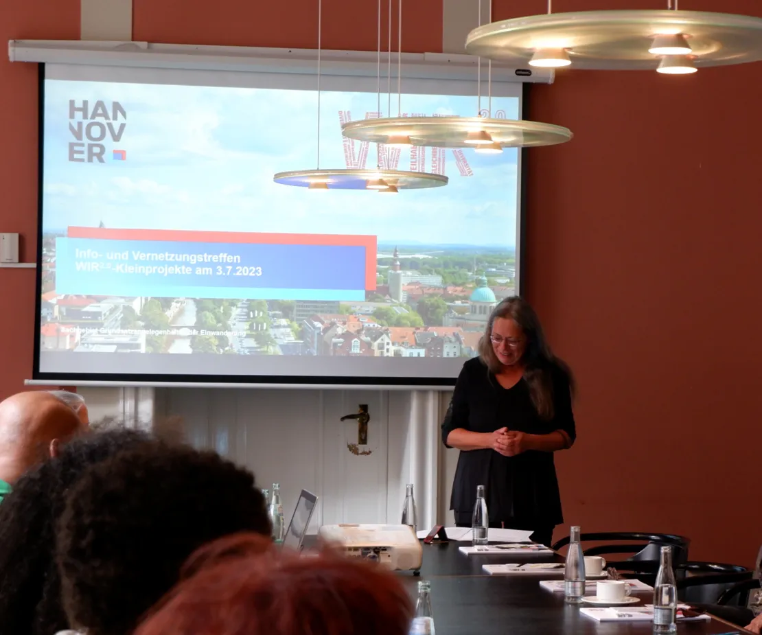 Eine Frau steht vor einer Leinwand. Auf der Leinwand die Begr&uuml;&szlig;ungsfolie einer Pr&auml;sentation. Im Hintergrund der Pr&auml;sentation eine Luftaufnahme von Hannover. Dar&uuml;ber der Titel "Info- und Vernetzungstreffen WIR2.0-Kleinprojekte am 3.7.2023".
