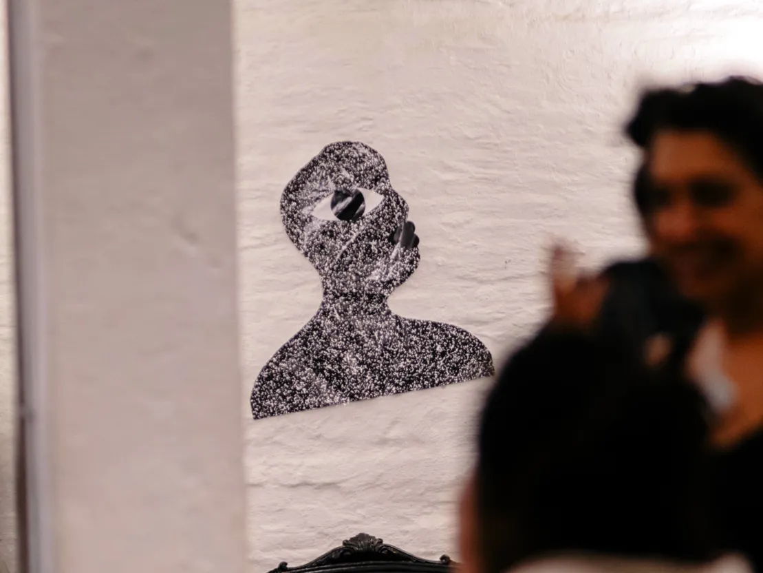 An einer wei&szlig;en Wand im Hintergrund klebt eine stilisierte, ausgeschnittene B&uuml;ste. Sie dreht den Kopf ins Profil, ein &uuml;bergro&szlig;es Auge prangt im Gesicht.