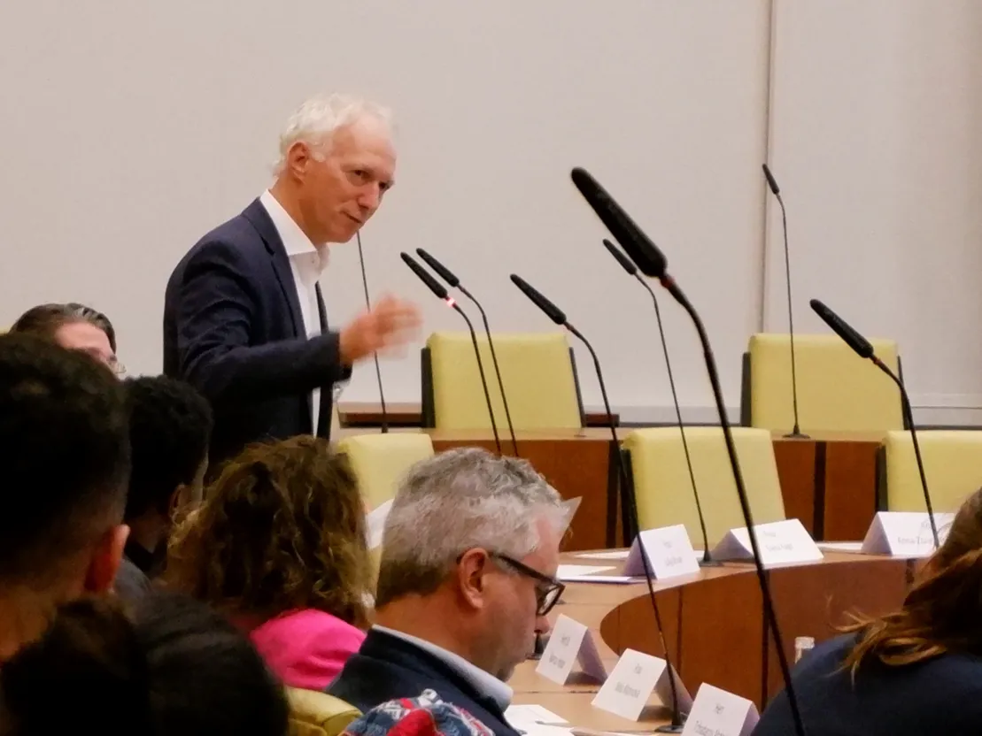 Ein Mann in dunkelblauem Anzu spricht in ein Tischmikrofon und gestikuliert mit der Hand. Im Bildvordergrund mehrer hinterk&ouml;pfe.