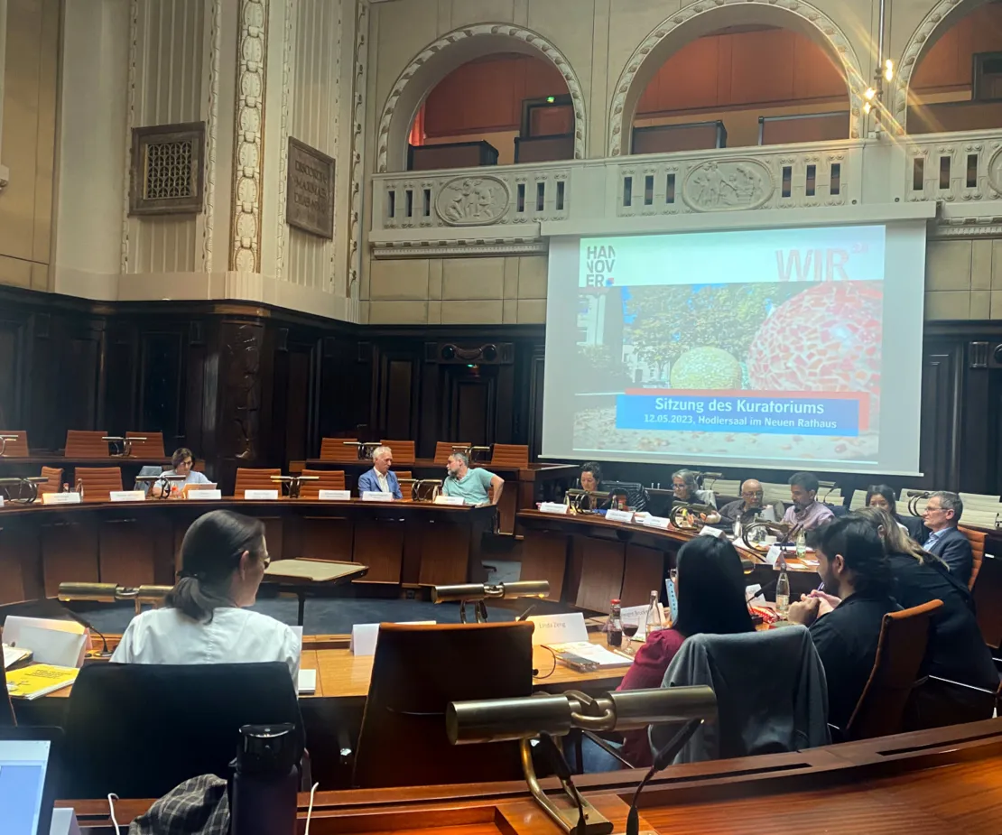 Eine Sitzung im Hodlersaal im Neuen Rathaus. Der Saal ist an den W&auml;nden mit dunklem Holz vert&auml;felt. Die fest installierten Holztische sind in zwei Reihen kreisrund angeordnet. An den Tischen sitzen &uuml;ber das Bild verteilt 13 Personen, die teilweise mit ihren NachbarInnen Diskutieren. Rechts im Hintegrund ist auf einer Leinwand eine Folie mit dem Text "WIR2.0 Sitzung des Kuratoriums" zu sehen.