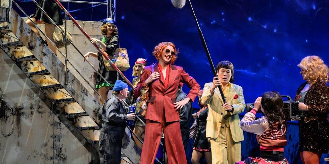 Schauspielende auf einer Bühne, die ein Kreuzfahrtschiff darstellen soll, stellen eine Pressekonferenz nach.