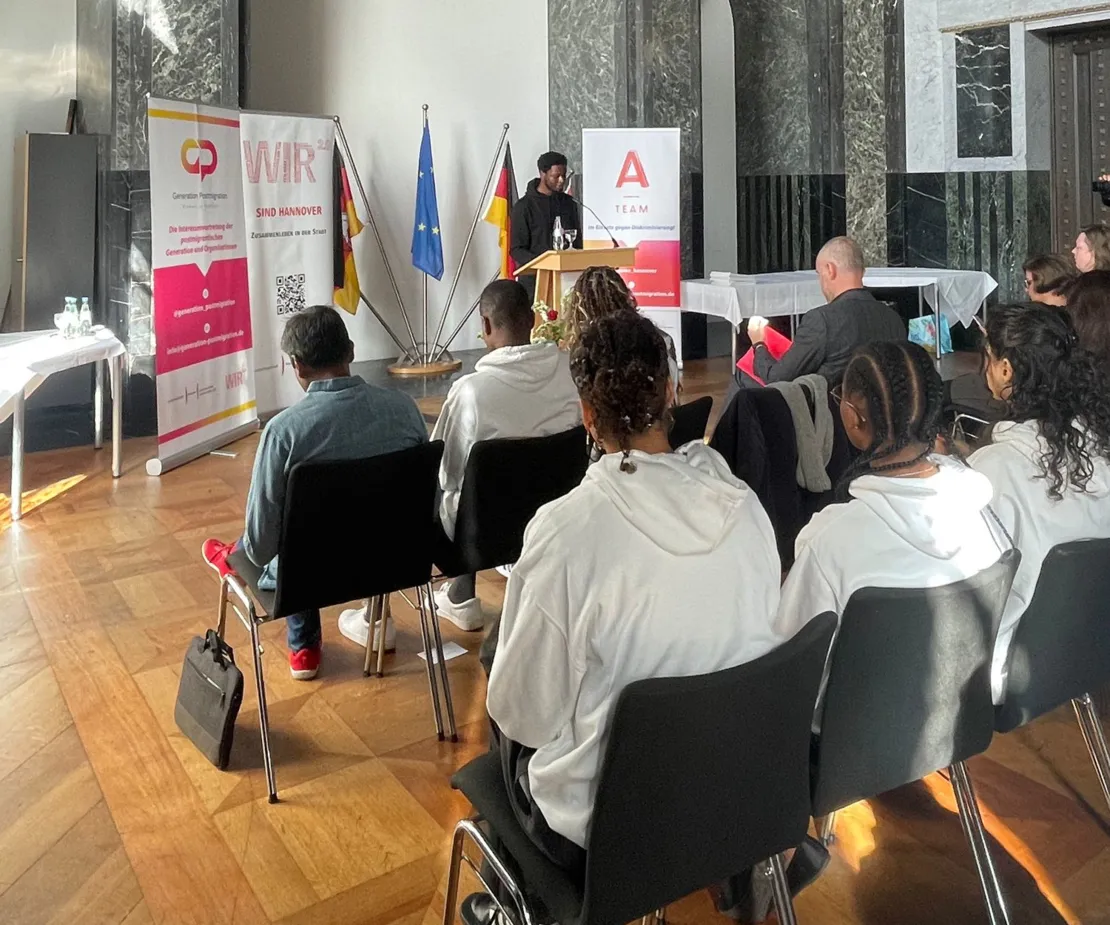 Tchadarou Abdoul spricht vor einigen jungen Menschen und OBn Onay. Neben ihm sind drei Banner zu sehen: Eines vom Netzwerk "generation Postmigration, eines vom WIR2.0-Prozess und eines von den A-Teams.