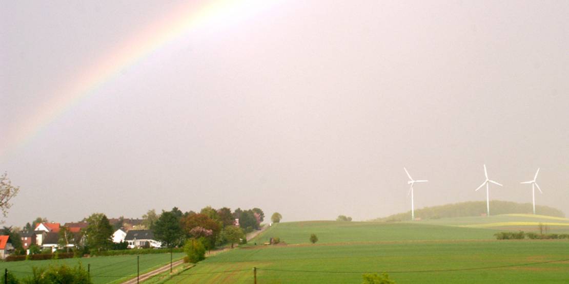 Drei Windräder stehen auf einem Hügel, etwas tiefer stehen mehrere Häuser. Am Himmel leuchtet ein Regenbogen.