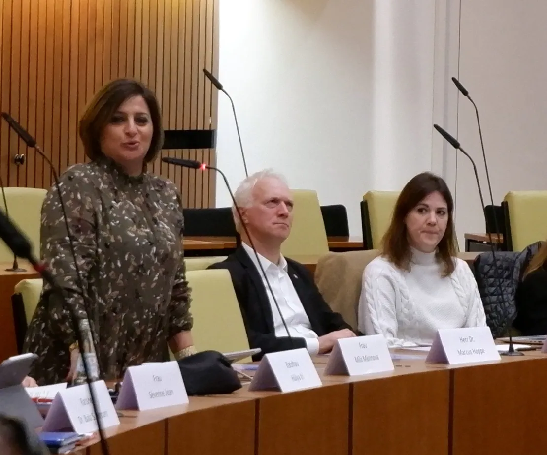 Eine Frau mit dunklen Haaren steht an einem Tisch und spricht in ein Mikrofon. Neben ihr sitzt ein Mann im schwarzen Sakko. Sein Gesicht dr&uuml;ckt keine Zustimmung aus.