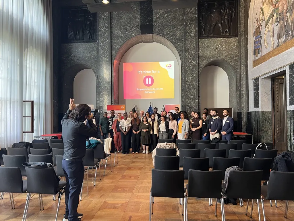 Eine Person in dunkler Kleidung fotografiert eine Gruppe von 25 Menschen in einem Saal.
