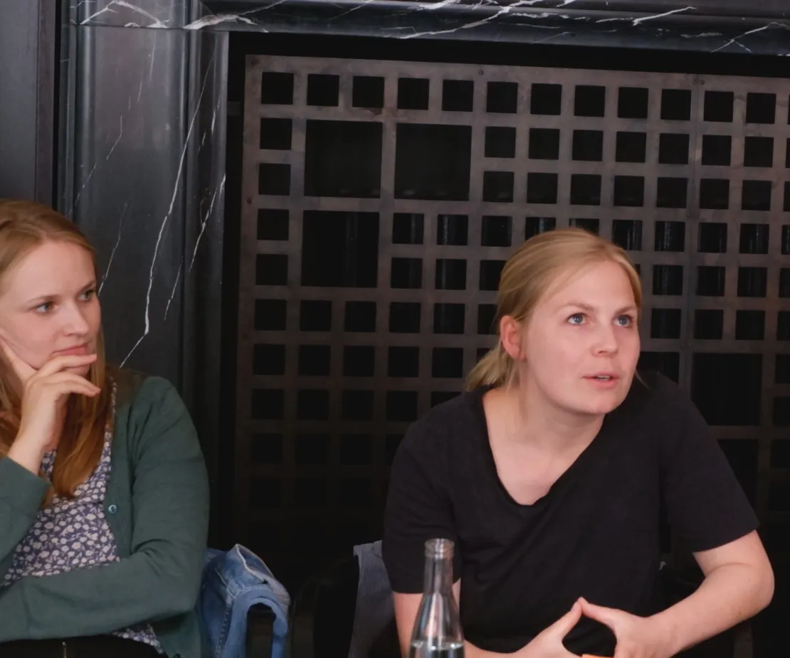 Zwei blonde Frauen sitzen an einem Tisch vor einem gro&szlig;en schwarzen Kamin.