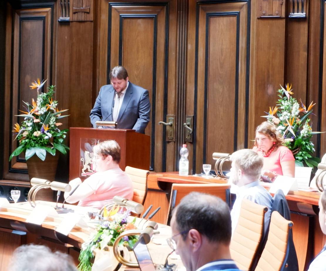 Ein Mann steht am Rede-Pult im Hodlersaal. Links und rechts neben dem Pult stehen dekorative Pflanzen. Im Vordergrund mehrere Menschen, die dem Mann zuhören.