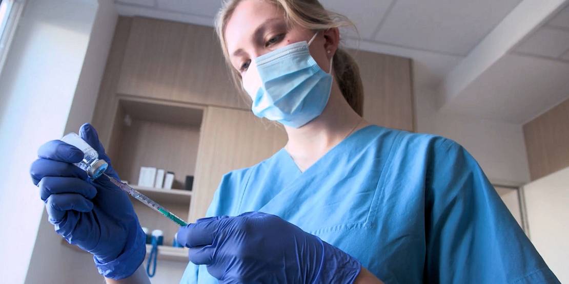 Frau mit Atemschutzmaske, blauem Kittel und Handschuhen zieht eine Spritze auf.