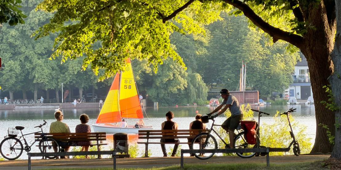 Maschsee: Vier Personen sitzen am Ufer auf Bänken und schauen auf ein Segelboot auf dem Maschsee.