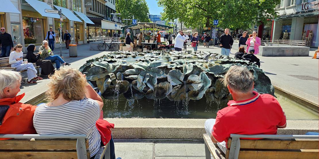 Menschen sitzen auf Bänken um den rechteckigen Blätterbrunnen herum.