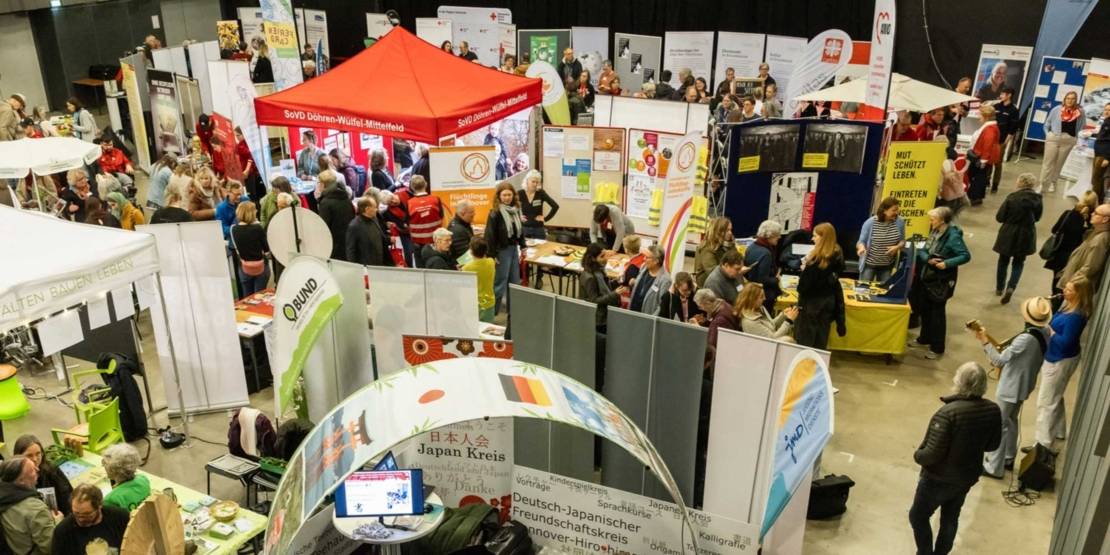 Blick von oben in einen großen Raum, in den sich viele Menschen zwischen Ständen tummeln. Rechts stehen zwei Musiker*innen.