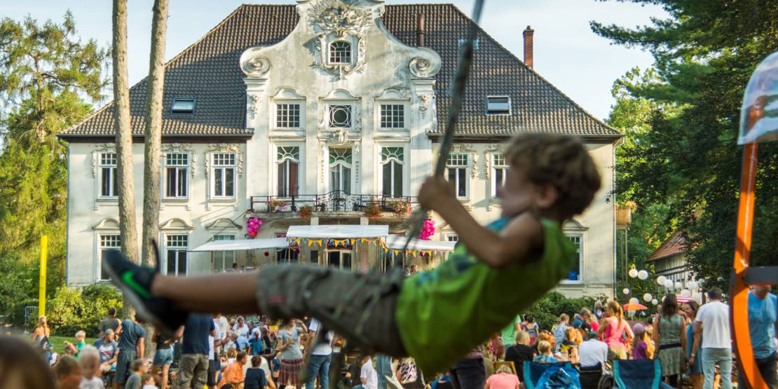 Ein Junge schaukelt. Im Hintergrund ist eine Rasenfläche vor einem Haus zu sehen, auf der sich viele Menschen befinden.