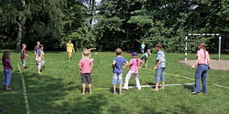 Auf einer Rasenfläche ist mit blau-gelbem Flatterband ein Spielfeld abgesteckt. Darauf spielen Kinder Völkerball: Die Kinder der gegnerischen Mannschaft werden mit einem Ball abgeworfen.