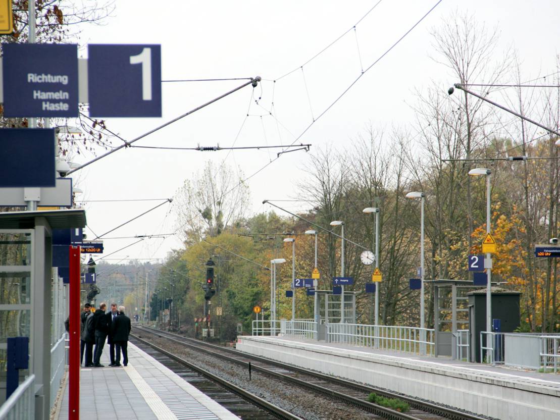 Blick auf den Bahnsteig des S-Bahnhofs Empelde. Links warten Menschen an Bahnsteig 1, rechts leuchtet ein rotes Signal an Bahnsteig 2, dazwischen verlaufen die Schienen.