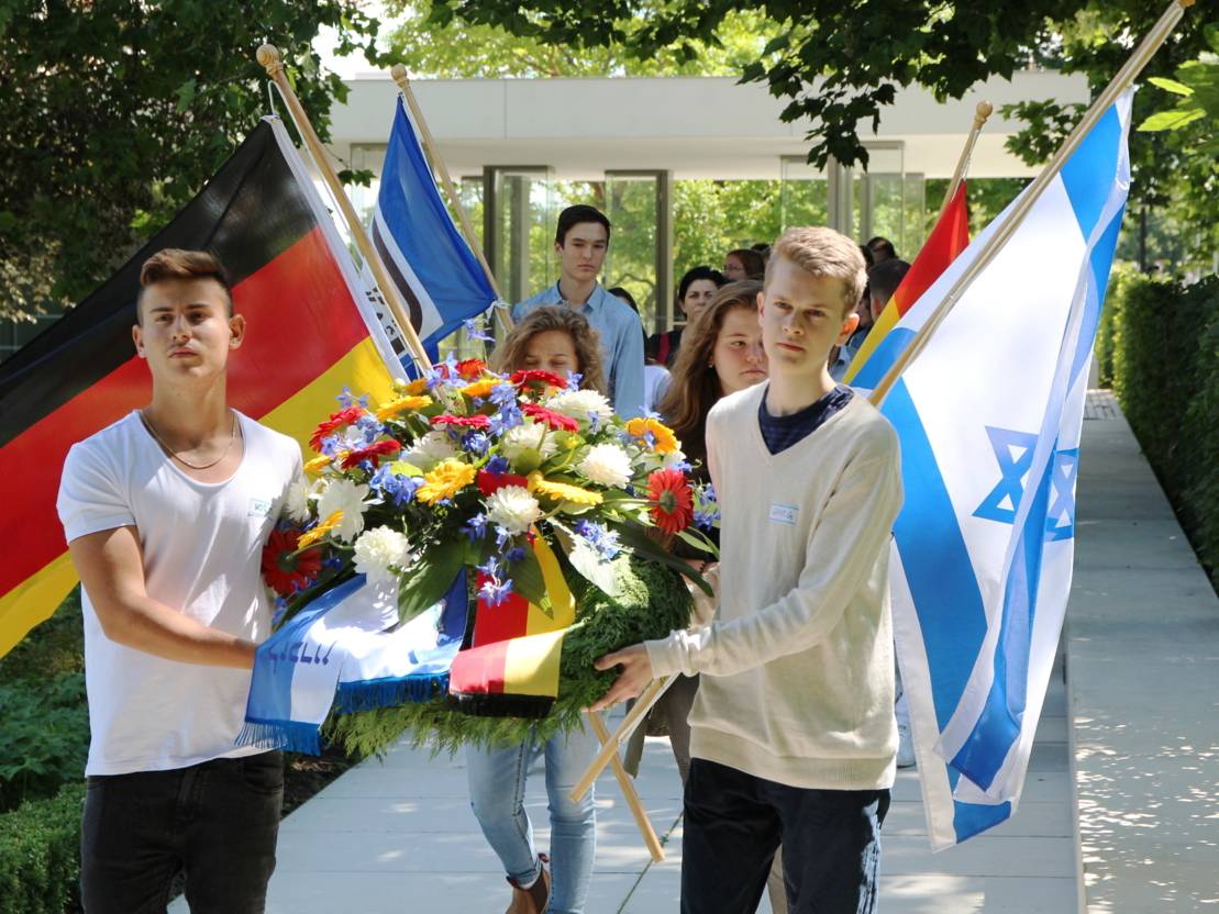 Zwei Jugendliche tragen einen mit Blumen geschmückten Kranz, dahinter gehen weitere Jungen und Mädchen mit Fahnen, zum Beispiel die des Staates Israel und der Bundesrepublik Deutschland.