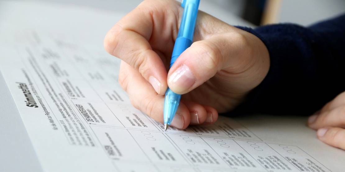 Eine Hand hält einen blauen Kugelschreiber und liegt auf einem Stimmzettel.