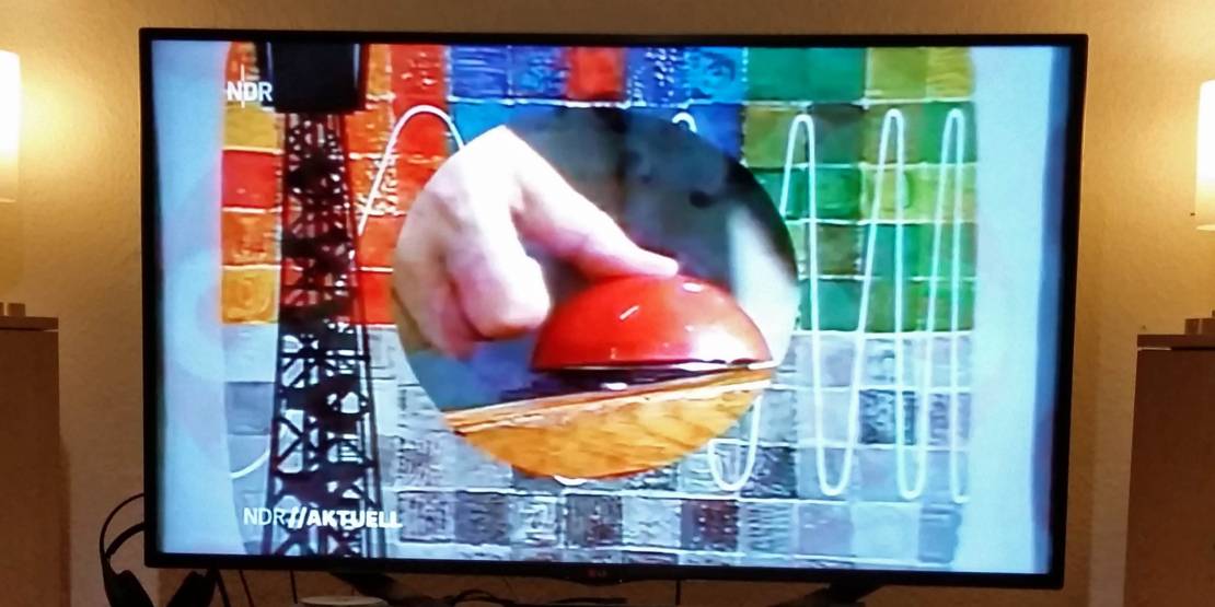 Fernsehbild in Farbe, das einen einen Daumen, der auf einen Knopf dr&uuml;ckt, zeigt