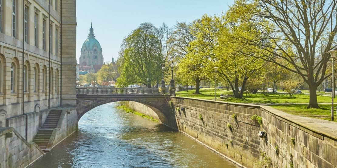 Fluß mit Gebäude am linken Ufer, Bäumen und Fußwegen am rechten Ufer, im Hintergrund ein Kuppelbau.