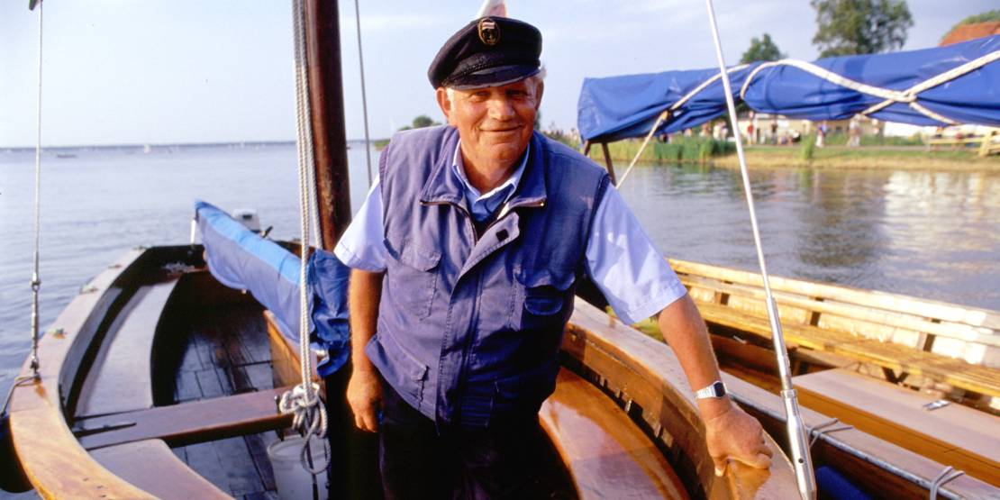 Ein Mann mit Schiffermütze und maritimer Kleidung steht in einem Boot des Typs "Auswanderer", einer offenen Segeljolle, welche als Ausflugsboot auf dem Steinhuder Meer unterwegs ist.