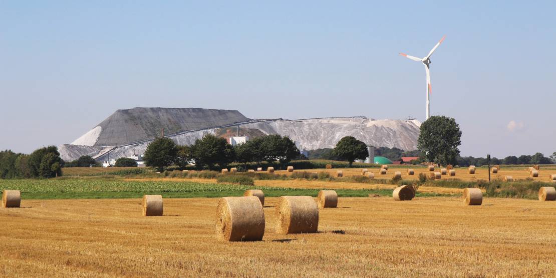 Stoppelfeld mit Strohballen, dahiner sind ein Kaliberg und Windräder zu sehen.
