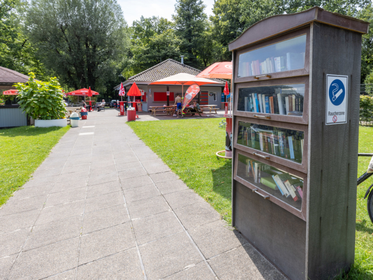 Bücherschrank an einem Plattenweg, im Hintergrund Sonnenschirme und Gebäude