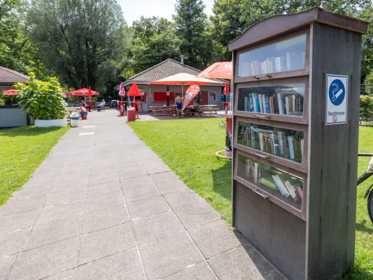 Bücherschrank an einem Plattenweg, im Hintergrund Sonnenschirme und Gebäude