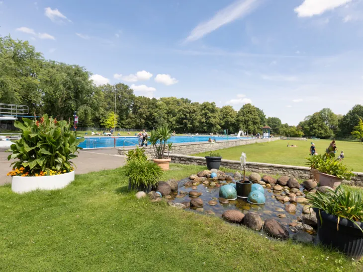 Springbrunnen, Rasenflächen, Kübelpflanzen, im Hintergrund ein Schwimmbecken