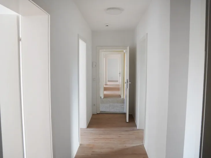 Blick durch einen langen hellen Flur, von dem Zimmer abgehen, in der Mitte vom anderen Boden eines Treppenhauses unterbrochen.