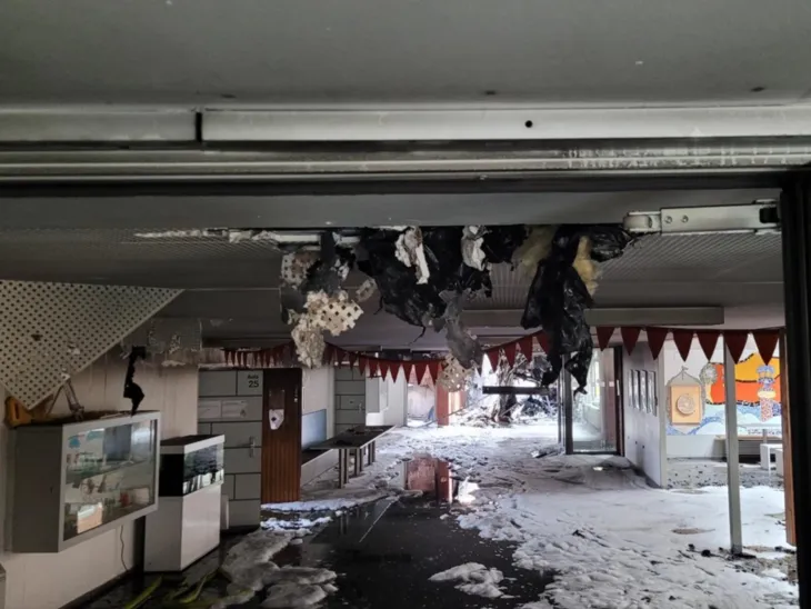 Blick in einen Gebäudeteil, der Boden mit weißem Schaum geflutet, von oben ranken schwarze Teile der Decke in den Raum. Nach links und rechts sind Gänge und Zugang zu enem breiteren Bereich zu erkennen. An den Wänden stehen und hängen Vitrinen, rechts ist eine Wand bunt gestaltet.